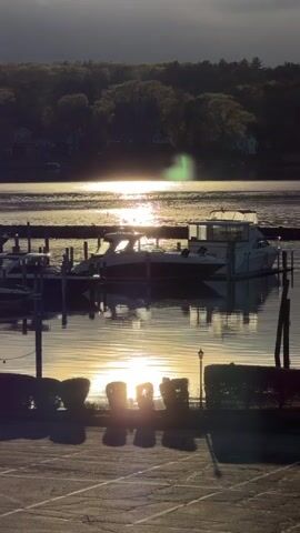 The video shows a boat dock scene in the late afternoon or early evening. A medium-sized motor yacht is docked at a wooden pier. The sun is low in the sky, reflected on the water, and slightly obscured by shrubbery in the foreground. Trees line the opposite shore, and a few houses are visible among them. A parking lot is in the foreground, with light post visible at the right edge. The sunlight creates strong reflections and some lens flare.