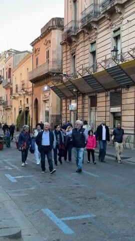 The camera pans across an urban street scene in what appears to be a European city. Many people are walking along the street, some in groups. Several are sitting at outdoor cafe tables, while one man is crouched down, possibly interacting with a dog. The architecture of the buildings is ornate, with balconies and detailed facades. The sky is light, suggesting it might be late afternoon or early evening. The street is paved and marked with blue squares, potentially indicating parking or pedestrian areas.
