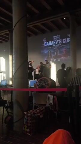 The video captures an arm wrestling match. Two men are seated opposite each other at an arm wrestling table, their hands clasped. A referee stands between them, wearing a striped shirt, and appears to be giving instructions or starting the match. In the background, a large screen displays text related to the event, including "KING OF TABLE PRESENTS - BABAEV'S" and a date of "JUNE 13TH 2026." There are other individuals visible, including a person holding a phone to record the event. The environment appears to be an indoor venue, possibly an event hall, with lighting focused on the arm wrestling table.