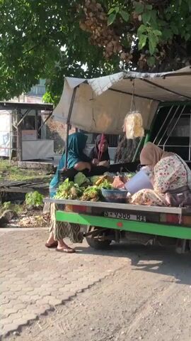 The video shows three women standing behind a pickup truck laden with fresh produce, suggesting a roadside market. The truck has a white canopy providing shade. One woman on the left, wearing a turquoise hijab and patterned pants, appears to be a customer or assistant, leaning forward. In the center, a woman in a pink hijab is interacting with someone, possibly a customer, and a pile of vegetables is visible in front of her. On the right, a woman in a beige hijab and floral-patterned top is seated and looking down, holding what appears to be a scale or a bag. The truck bed is filled with various vegetables and other goods, including what looks like dried snacks hanging from the canopy. The ground is paved with interlocking bricks, and a tree with green foliage casts shadows over the scene. In the background, a building with white walls and a tiled roof is partially visible. The overall impression is a bustling, everyday market scene under a bright sky.