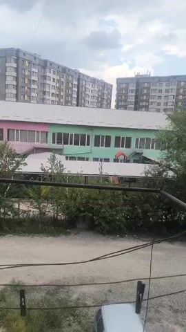 The video shows a view of a street with several buildings and trees. In the foreground, there are power lines and a section of a white car. Behind the power lines, there are trees and bushes. Further back, there is a long, low-rise building with a light-colored roof and walls painted pink and green, with white-framed windows. Behind this building, there are two tall, multi-story apartment buildings with balconies and many windows. The sky is overcast with some clouds.