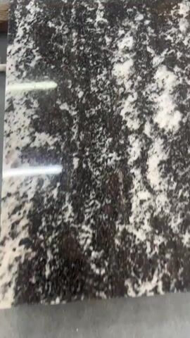 This media file captures a close-up, angled view of a granite slab. The granite has a dark base speckled with white and some reddish-brown flecks, creating a mottled, natural pattern. The surface appears polished, as evidenced by the bright reflections of overhead lighting, which streak across the stone. The camera moves slowly, offering a slightly shifting perspective of the granite's texture and variation. No people, text, or specific environmental cues are visible, making it difficult to determine the exact setting beyond it being a location where granite slabs are present, such as a showroom, workshop, or fabrication facility. The lighting suggests an indoor environment.