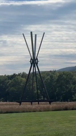 The video shows a panoramic view of a park-like landscape. In the foreground, there is a well-maintained green lawn. Behind the lawn, a field of tall, dry grass stretches across the middle ground. In the background, a dense forest of green trees forms a backdrop, with hills visible beyond the trees under a partly cloudy sky. Two large metal sculptures are visible in the scene. The first sculpture, on the left side of the frame, is tall and has a tripod-like base with slender metallic poles extending upwards and outwards. The second sculpture, further to the right and partially obscured by the grass, is red and appears to be abstract with multiple angled limbs. The camera pans slowly from left to right, capturing the expanse of the landscape and the sculptures within it. There are no people visible in the video.