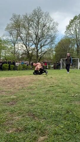 Two shirtless men wrestle on a grassy field in front of a crowd of onlookers. One man is on top of the other, grappling for dominance. A woman stands nearby, seemingly observing the match. A soccer goal is visible on the edge of the field. Trees surround the field, and the sky is overcast.