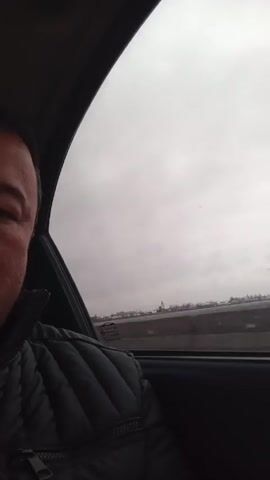 The video, shot from inside a moving car, shows a roadside view through the passenger window. The sky is overcast. A highway with a concrete barrier runs parallel to the car's movement. Trees, blurred due to the speed, pass by the window. The partial view of a person wearing a quilted black jacket is visible in the lower left corner of the frame.