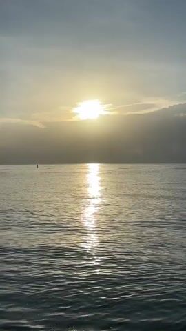 The video captures a serene seascape at sunrise or sunset. The sun is partially obscured by clouds, casting a bright, shimmering reflection across the calm water. The horizon is visible in the distance, with the sky a pale gradient of blue and yellow. The water is rippled, suggesting a gentle breeze. There are no people or discernible objects in the water, besides a very small, distant marker or buoy. The overall mood is peaceful and tranquil.