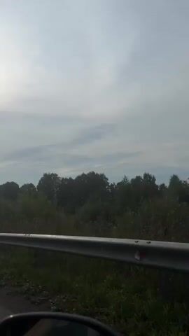The video shows a car's perspective as it travels down a road. On the left side of the road is a metal guardrail, and beyond that is a grassy field with trees and vegetation. In the distance, there are a few houses or buildings. On the right side of the road, there are more trees and a utility pole with power lines. The sky is cloudy, and the sun is visible, casting a bright light. The video captures a rural or semi-rural landscape.