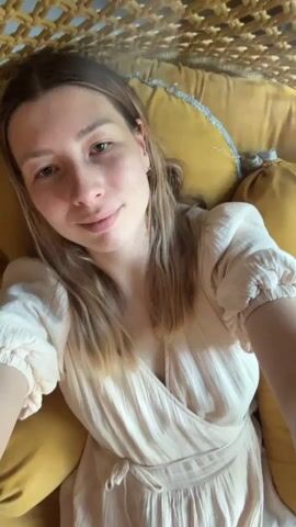 A woman with blonde hair and light skin is laying on a yellow pillow. She is wearing a light pink dress and looks directly at the camera with a slight smile. The background consists of a woven rope-like structure. A baby coos in the background.