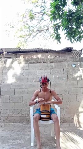 A shirtless young man with braids and glasses, wearing a multi-colored helmet, is sitting in a white plastic chair in front of a cinder block wall. He is playing a small drum placed between his legs. Above him, a tree with leaves extends into the frame from the top right corner, and another tree branch with fewer leaves is visible in the upper left corner. The ground appears to be a mixture of dirt and concrete. A section of corrugated roofing is attached to the top of the cinder block wall.