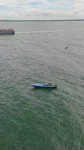 An aerial view of a body of water shows a large barge with a blue hull moving from left to right across the frame. In the foreground, a smaller blue boat with a white top and orange flags is positioned to the right of the frame, moving slightly towards the top right. A white object, possibly a bird, is visible in the water to the right of the small boat. The sky is overcast with clouds.