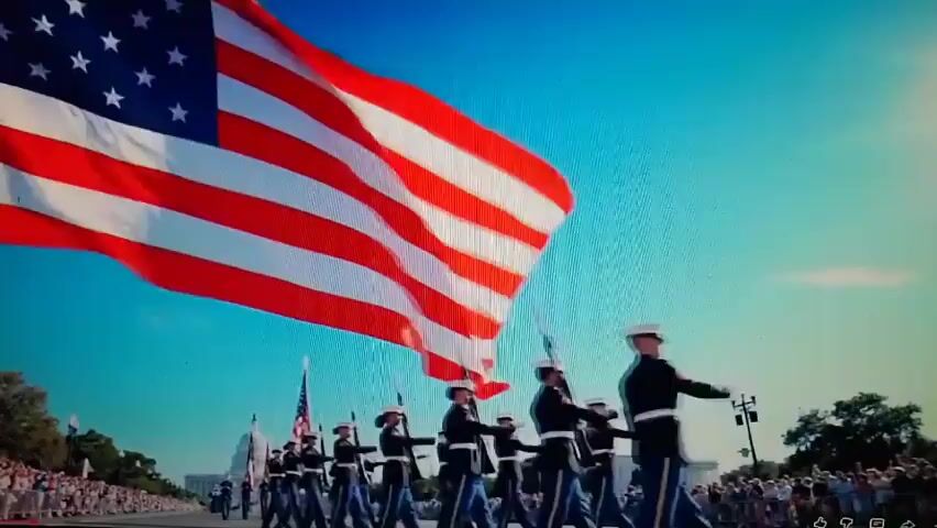 The video begins with a low-angle shot of a large American flag waving against a bright blue sky. A procession of soldiers in uniform, carrying rifles, marches in the foreground. In the background, the Washington Monument and the U.S. Capitol Building are visible, along with a crowd of spectators. The scene appears to be a parade or ceremony.

The second part of the video shows a parade float adorned with red, white, and blue flowers and an eagle statue. The words "America 250" are visible on the float. Soldiers with rifles and flags are marching in front of the float, and a crowd of spectators is gathered on the sidewalks, some holding up phones to take pictures. The buildings in the background appear to be in a city. The overall impression is a patriotic celebration.