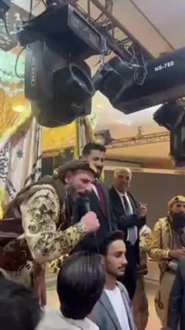 A group of men are celebrating in what appears to be a banquet hall. Several are wearing traditional headwear and garb. A man with a microphone is speaking.  Others are clapping and cheering. There are light fixtures and decor on the ceiling. The room is brightly lit.