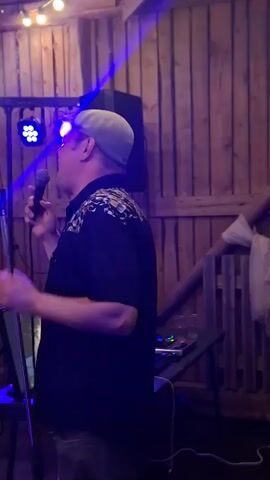 A man wearing a flat cap and a patterned shirt is holding a microphone and singing into it. He is standing in front of a wooden wall. There are stage lights in the background, casting colored light on the scene. A speaker and a table with electronic equipment are also visible.