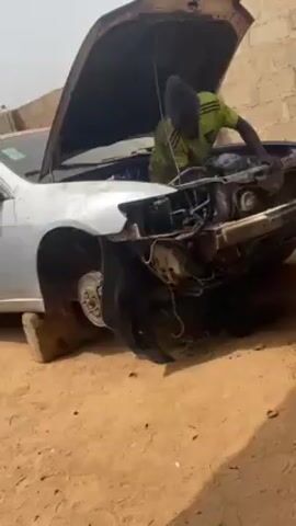 The video shows a man working on a white car outdoors. The car's hood is open, and the front bumper is missing. The car is raised on blocks. The man is wearing a neon green shirt and a hat, and he is bent over inside the engine compartment. The surrounding area is dirt and there's a brick wall nearby. The video is shot from a low angle.