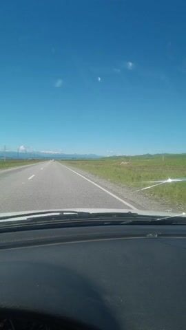 This is a first-person point-of-view video taken from inside a car. The camera is pointed forward, showing the road ahead and the sky. The car is driving on a paved road that appears to be a highway or a major country road. The road is bordered by green fields and distant mountains. The sky is clear and blue with a few scattered white clouds. The dashboard and steering wheel of the car are visible in the foreground, indicating that someone is driving. The video captures the sensation of traveling on an open road under a bright, sunny sky.