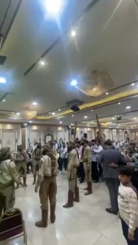 A group of men in traditional clothing are dancing in a large, ornate hall. The men are wearing tan and white outfits with intricate embroidery and head wraps. Some of them are carrying flags. The hall is filled with people who are watching and taking photos. The room is decorated with chandeliers and other luxurious furnishings. The lighting is bright.