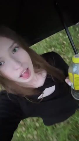 This media file captures a person holding an open black umbrella and a yellow-capped bottle outdoors. The person, with long brown hair, is wearing a black shirt and appears to be in a natural setting with green grass visible. The scene is blurred due to motion, suggesting the camera is moving or the person is. The weather appears to be overcast, with a pale sky visible through the trees. The specific location is Nevyansk, Russia. The overall mood is casual and perhaps a bit playful, given the dynamic camera movement and the person's expression. No visible text is present in the media file.
