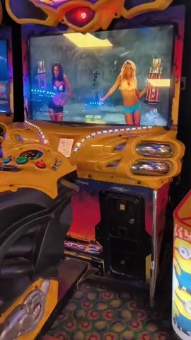 The video shows an arcade. In the foreground, there are two arcade machines. The machine on the left is a motorcycle racing game with a yellow and black seat and handlebars, and a yellow controller. The screen shows a wrestling game with two women. The machine on the right is a yellow and red Minions-themed game. In the background, more arcade machines are visible, some with bright lights. The floor is covered in a patterned carpet.