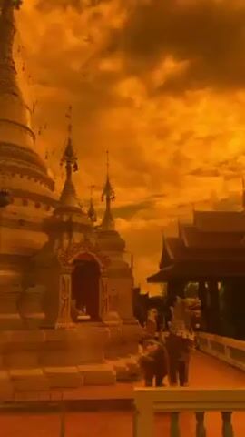 The video shows a Buddhist temple, likely in Southeast Asia, under an orange-colored sky with clouds. The temple is made of golden colored materials and has pointed roofs. There are statues of lions. The lighting suggests either sunset or artificial coloring. The overall impression is that of a foreign, cultural landscape.