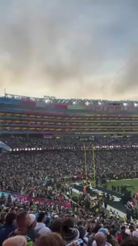 A large crowd fills a football stadium on an overcast day. On the field, a group stands on a decorated platform while music plays. The stadium’s upper deck is visible, with advertising lining the top. The camera captures the atmosphere from the perspective of someone in the stands.