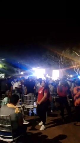 The video depicts a nighttime outdoor gathering. Several people are walking around, seemingly at a party or event with music and bright lights in the background. Many of the attendees are dressed in casual clothes. Some chairs are visible, suggesting seating arrangements. There is general movement and ambient sound, typical of a social gathering.