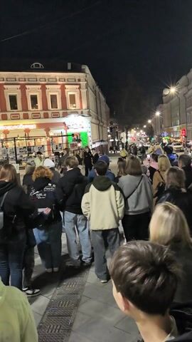 The video shows a street scene at night, crowded with people. Many people are wearing jackets and casual clothing. The street is brightly lit by streetlights and lights from nearby businesses.  A building with a red and white facade is visible in the background. Some of the people are looking in the same direction as the camera. Some are interacting with others nearby. The overall impression is that there is a gathering or event taking place in the street.