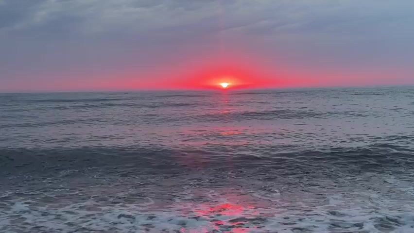 The video shows a sunset over the ocean. The sun is a bright red orb on the horizon, casting a pink glow over the clouds and the water. Waves crash on a pebble beach in the foreground. The scene is peaceful and serene.