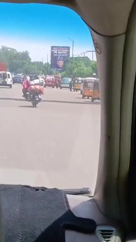 The video shows a view from inside a car on a road with traffic. The traffic includes cars, motorcycles, and auto-rickshaws (tuk-tuks). A billboard is visible in the distance. The sky is blue and the weather appears sunny. The interior of the car is visible in the foreground.