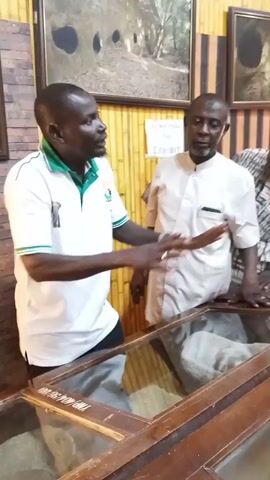 The video shows four African men inside a museum. They are standing around a glass display case, and the man in the white polo shirt is gesturing with his hands. The man in the white tunic and another man in a patterned shirt lean on the case.  A fourth man wearing a green baseball cap and white Adidas jacket stands nearby holding a bottle of water.  The walls are paneled with bamboo and feature framed photographs.  A sign is visible that reads "DO NOT TOUCH THE EXHIBIT."