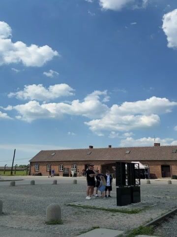 The media displays a series of short clips showing the grounds of a historical site, likely Auschwitz-Birkenau in Brzezinka, Poland. The subjects include several people, some of whom appear to be tourists or visitors, observing the surroundings. Objects visible include brick buildings with red tile roofs, a gatehouse with a tower, train tracks, and some modern art installations or informational displays. The scene depicts the exterior of barracks-like structures and open, grassy areas.

The activity taking place is general observation and exploration of the site. There are no overt actions indicating a specific event.

Notable details include the clear blue sky with scattered white clouds, suggesting fair weather, and what appears to be midday given the lighting. The architecture of the buildings, the train tracks, and the overall layout strongly indicate a former concentration camp complex. The presence of visitors suggests it is now a memorial or museum. The atmosphere conveyed is one of somber reflection, though this is interpreted through the context of the location rather than explicit emotional cues from the individuals. No visible text is present in the clips.