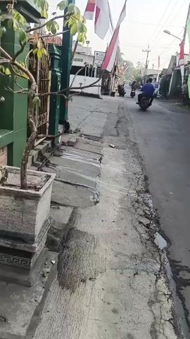 The video shows a street with several motorcycles. On the right side of the street, there is a building with a green wall and text that says "PONDOK PESANTREN DARUL FALAH 6 WATESARI". There are also Indonesian flags and banners on poles along the street. The road appears to be paved and in fair condition, with some cracks and patches. There are trees and bushes planted along the side of the building. The lighting suggests it is daytime, possibly morning or afternoon. One person is visible riding a motorcycle in the distance.