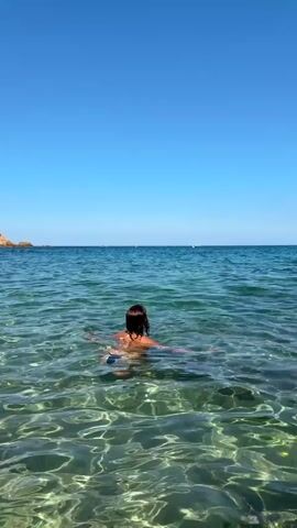 A person in a black bikini jumps into clear, shallow water, creating a splash. The person is facing away from the camera, with arms raised. The water is calm and the sky is clear blue. The scene appears to be a beach or a coastal area.