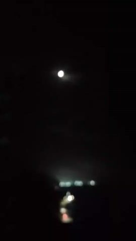 The video shows a dark night scene. A bright moon is visible in the upper center of the frame. Below the moon, a line of blurred lights suggests a distant town or settlement. The lights are scattered and appear to be along a road or a body of water. The foreground is dark, with some indistinct shapes that could be trees or foliage on the left side. The overall impression is a nighttime landscape with a moonlit sky and distant human activity.