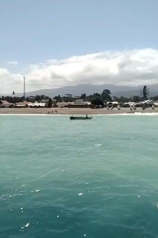 The video shows a wide shot of a coastal village scene. In the foreground, the turquoise water of the ocean is visible with some whitecaps. A thin, light-colored line, likely a fishing line or rope, stretches across the water. In the midground, a small, dark boat with at least one person in it is moving on the water. Behind the boat is a sandy beach with several structures, including houses and what appears to be a communication tower. A line of people are visible walking along the beach. Further in the background, lush green mountains rise under a blue sky with scattered white clouds. The overall impression is a peaceful, tropical coastal setting.