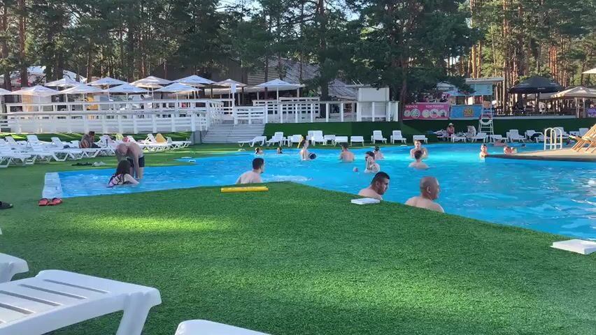 A wide shot shows a busy outdoor swimming pool area on a sunny day. The pool itself is a vibrant blue, surrounded by artificial green turf and white lounge chairs. Several people are in the water, some swimming, others standing and socializing. In the foreground, a group of men are visible in the pool, with one man leaning against the edge. To the left, a woman with dark hair is partially submerged in the water, and a yellow pool noodle floats nearby. Further in the background, there are more people lounging on chairs under white umbrellas and several buildings with awnings. The area is bordered by a dense forest of tall, green pine trees. The overall atmosphere appears to be one of leisure and recreation.