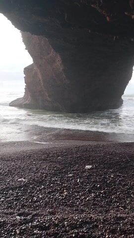 The video shows a rocky beach from the perspective of someone standing under a rock formation. The beach is covered in small, dark pebbles. In the distance, a large rock formation stands in the ocean, with waves gently lapping at its base. The sky is overcast, providing soft, diffused light. There are no people visible in the scene, and the focus is solely on the natural elements of the beach and rock formations.