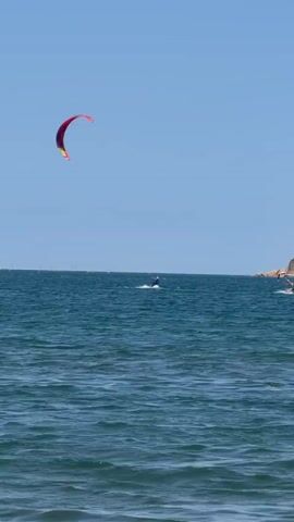 A kitesurfer is performing a jump with a red and yellow kite over the choppy blue ocean. In the distance, another kitesurfer is visible closer to a wooded island with buildings and a lighthouse on its shore, under a clear blue sky.