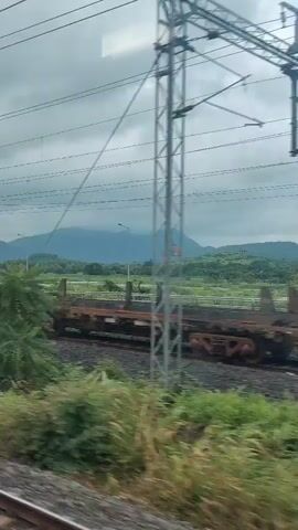 A train passes by a rural landscape with green hills in the background. Overhead power lines for the train are visible. In the foreground, there are railway tracks and lush green vegetation. A few buildings are sparsely scattered in the distance. The sky is overcast.