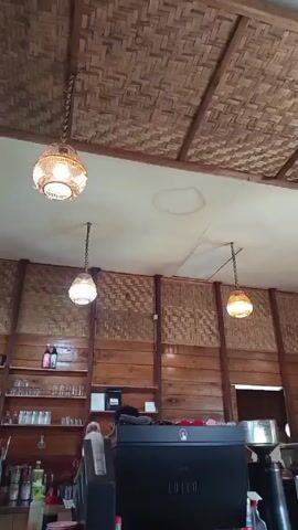 The camera pans left across the interior of a cafe or restaurant. The ceiling is comprised of woven bamboo and hanging spherical wicker light fixtures. The wall is wood paneling with woven bamboo sections. The cafe counter is also made of wood. Shelves hold glassware and bottles. A refrigerator is visible. A black "LOLLO" espresso machine stands on the counter. A person is behind the counter.