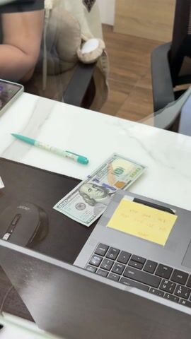 In this video, a woman behind a counter is counting Thai Baht and some coins, placing them next to a $100 bill. The setting appears to be a money exchange booth, as indicated by the signage, "Money Changer" on a receipt. The desk has a laptop, a mouse, a pen, and a phone with an image on the screen. The woman is wearing a black t-shirt with the number 24 on it, a gold bracelet, and has manicured nails.