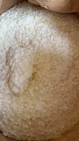 A close-up shot shows a hand reaching into a bag of small white pellets. The hand scoops up a handful of the pellets, and they are shown in the palm and fingers of the hand. The pellets are uniform in size and shape, and they appear to be made of a waxy substance. The background is a blurred brown paper bag.