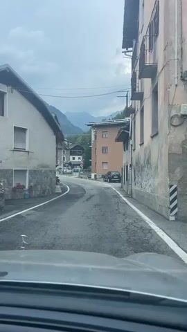 The video shows a view from inside a vehicle driving on a paved road through a small town. The road is winding and has some cracks in the pavement. On either side of the road are buildings, some made of stone and others with stucco exteriors. In the background, mountains can be seen, covered in trees and under a cloudy sky. There are a few cars parked along the road and one moving vehicle in the distance. Traffic signs are visible, including a speed limit sign. The architecture of the buildings suggests a European setting.