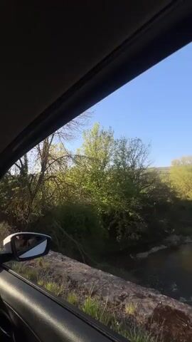The video shows a view from a vehicle, likely a car, overlooking a small river or stream in a wooded area. The camera is positioned at a slight angle, capturing the river flowing below a stone ledge. Lush green trees and vegetation line the riverbanks. The sky above is clear and blue. There is the sound of someone speaking in the background, but the audio is muffled and hard to discern the content.