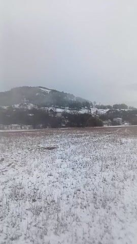 The video captures a cold, overcast winter scene in an open field covered in patchy snow. Distant hills or mountains, partially obscured by mist or low clouds, form the backdrop. Some trees and scattered buildings are visible at the base of these hills.

At the 0:01 mark, a person enters the frame from the left, walking slowly across the snow-covered field towards the right. They are dressed in winter camouflage attire, including a jacket and pants, black gloves, and a black beanie with a red emblem on it. A red backpack is slung over their shoulders. The person appears to be smoking, holding a small white object, possibly a cigarette, between their fingers near their mouth as they walk. They take a puff from the object.

As the person walks past the camera, two metal detectors are visible lying on the snow-covered ground in the foreground. One is predominantly grey and black with a prominent orange search coil, while the other is mostly black. The person glances back briefly before exiting the frame. The video concludes with a final shot of the snowy field and the distant, misty landscape under the grey sky. Patches of green are visible in some distant fields, contrasting with the white snow.