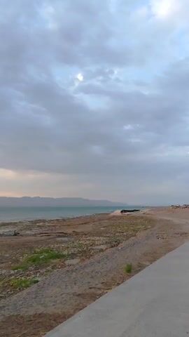 The video shows a shoreline with a body of water and mountains in the distance. The sky is cloudy. There is a road on the right side of the frame and the shoreline is on the left. There is a small boat in the water. The overall impression is of a calm, natural scene.