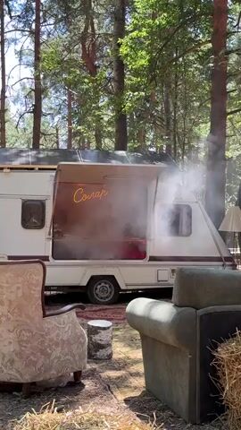 A retro-style camper trailer is parked in a wooded area. A neon sign that reads "Солар" is visible above the trailer's serving window. Smoke or steam is coming from the window. Two vintage armchairs are placed in front of the trailer, with hay bales on either side of the right armchair. A small tree stump is positioned between the armchairs. The ground is covered with dirt and dry leaves. Sunlight filters through the trees, casting shadows.