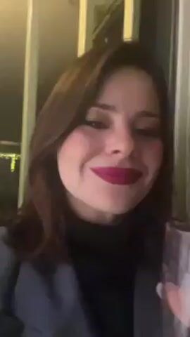 A woman drinks from a clear glass, presumably containing champagne or sparkling wine. She has shoulder-length brown hair, wears a dark top and bright pink lipstick. She smiles slightly before taking a sip.  The background suggests she may be outdoors or on a balcony with a light-colored railing. The video is shot close-up with the woman's face being the primary focus.