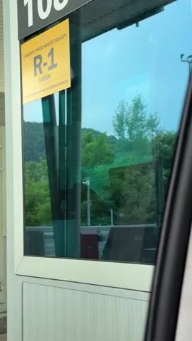 This is a medium shot video, filmed from a car, of a toll booth window. The window is tinted blue and reflects the surrounding trees and sky. On the window, a yellow sign is visible with text in what appears to be Bosnian. The text includes "U OBJEKTU PORED MOŽETE PREUZETI R-1 RAČUN" and an email address "ali po zahtjev u mail: acc@jpautoceste.ba". This indicates that receipts (R-1 RAČUN) can be obtained at the facility and can be requested via email. Inside the toll booth, two vertical green poles are visible, along with a red box and a dark monitor. The scene suggests a toll collection point on a road, likely in a rural or forested area, given the lush green trees and hillside in the reflection. The lighting suggests it is daytime, possibly overcast. The perspective from inside a vehicle implies the user is either approaching or has just passed through the toll.