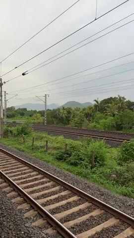 The video begins with a view from a train's window, showing train tracks and lush green vegetation. Mountains are visible in the distance under a cloudy sky. The perspective shifts to show the side of a red train car moving alongside the tracks. Next, the camera is inside a train car, looking towards a door and electrical equipment. Outside the open door, a rough, rocky terrain and more green vegetation are visible. The final shot is a blurry view of the landscape from the open door, with a rough road-like path and green hills in the background.