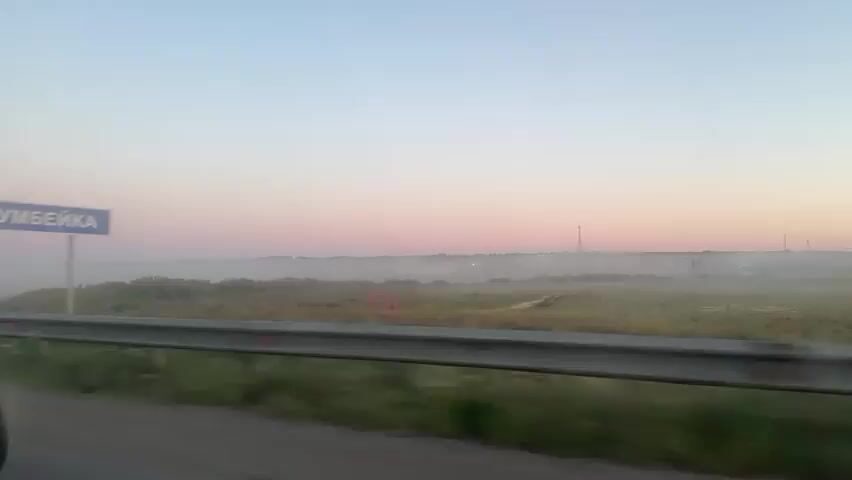 The video captures a view from inside a moving vehicle during what appears to be sunrise or sunset. The sky displays soft hues of pink and blue, with a gentle haze or fog settling over the landscape in the distance. A road barrier with reflective markers runs alongside the vehicle. In the first shot, a blurred blue sign with white text is visible on the right side of the road. The subsequent shots show the road stretching ahead, with a red-railed bridge or overpass in the middle distance. The interior of the car is partially visible, including the dashboard with a small metallic object on it and a portion of the windshield. The reflection of the sky and landscape can be seen in the car's side mirror in the second shot.